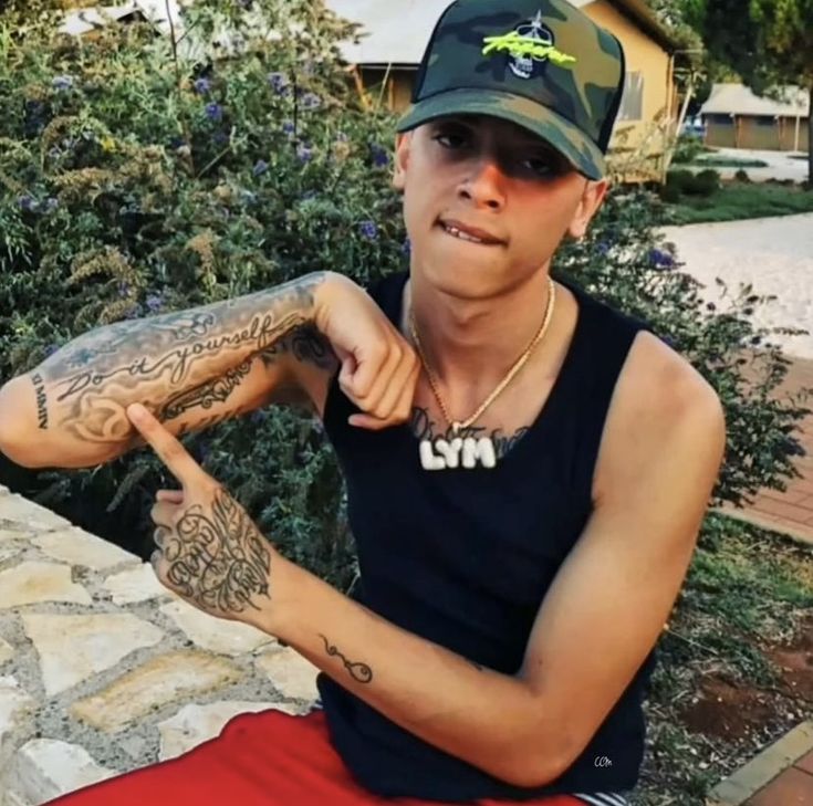 a man with tattoos sitting on a stone wall and pointing at the camera while wearing a hat