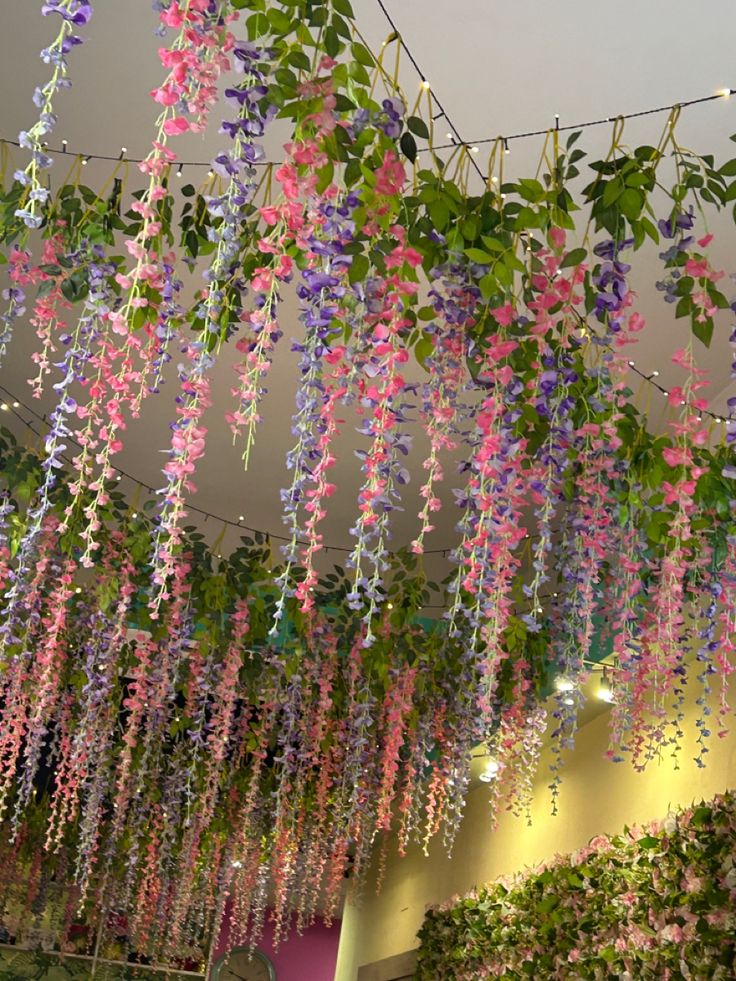 purple and pink flowers hanging from the ceiling in an indoor space with plants on either side