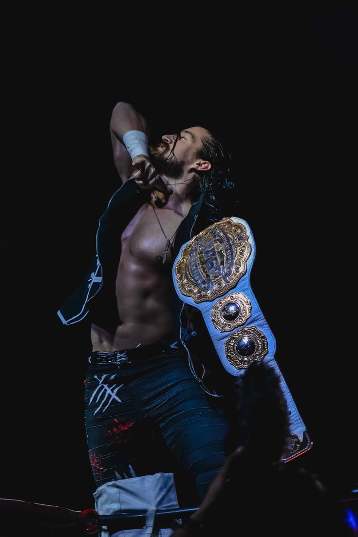 a shirtless man holding a blue and gold boxing belt in his right hand while singing into a microphone