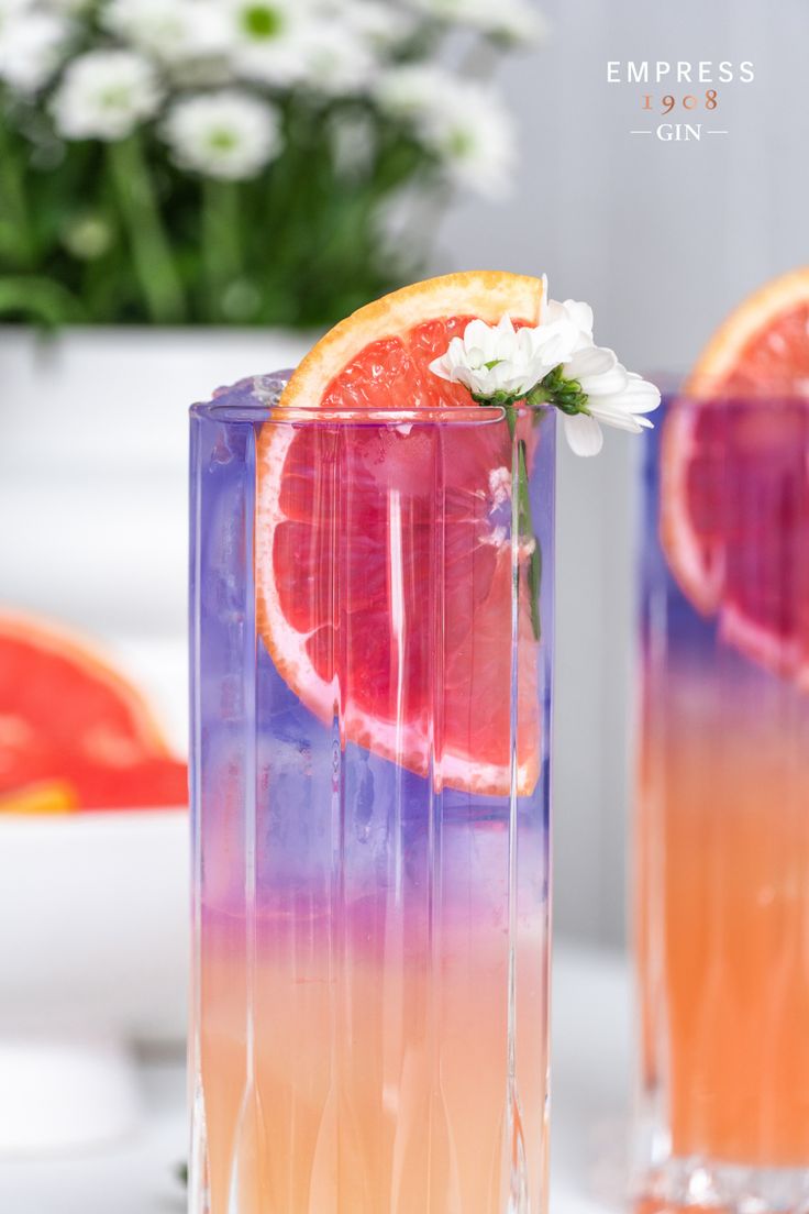 two glasses filled with orange slices and flowers on top of a white table next to watermelon slices