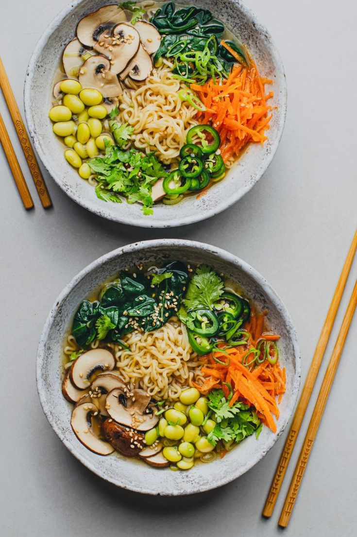 two bowls filled with noodles and vegetables next to chopsticks on a white surface