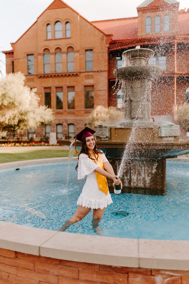 a woman in a graduation cap and gown standing next to a fountain with water spouting from it