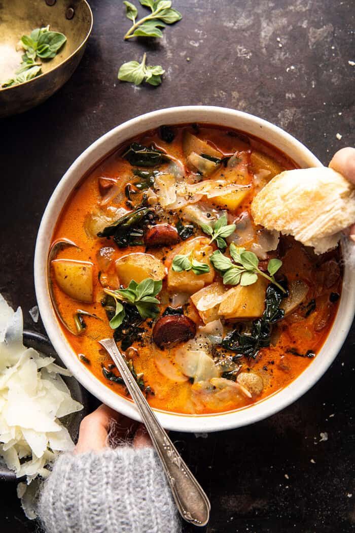 a person holding a spoon over a bowl of soup with potatoes and spinach on the side