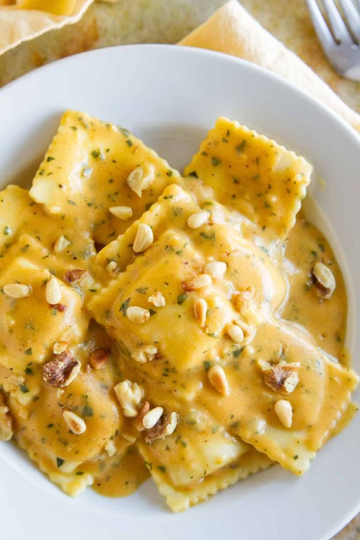 a white plate topped with ravioli covered in cheese sauce and nuts next to a fork