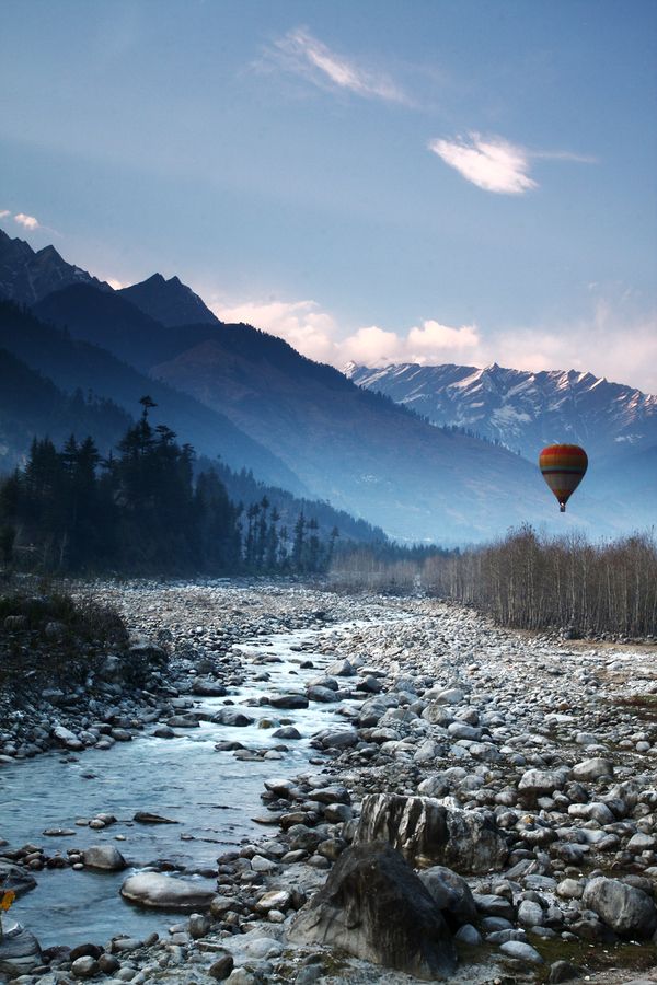 a hot air balloon flying over a rocky river with mountains in the backgroud