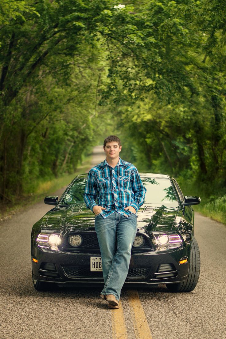 a man sitting on the hood of a black car in front of some green trees