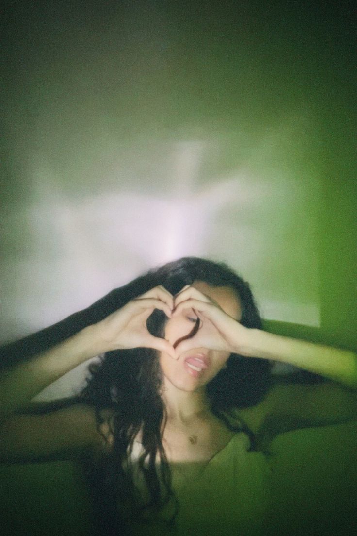 a woman making a heart with her hands while standing in front of a green wall