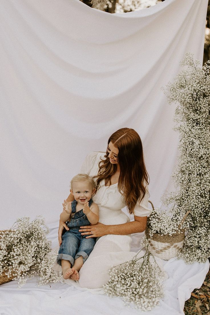 a woman holding a baby in her arms while sitting on the ground with flowers and greenery