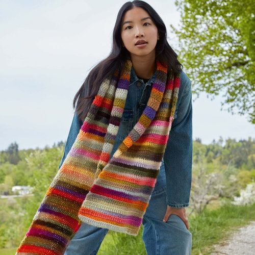 a woman standing in the grass wearing a multicolored knitted scarf and jeans