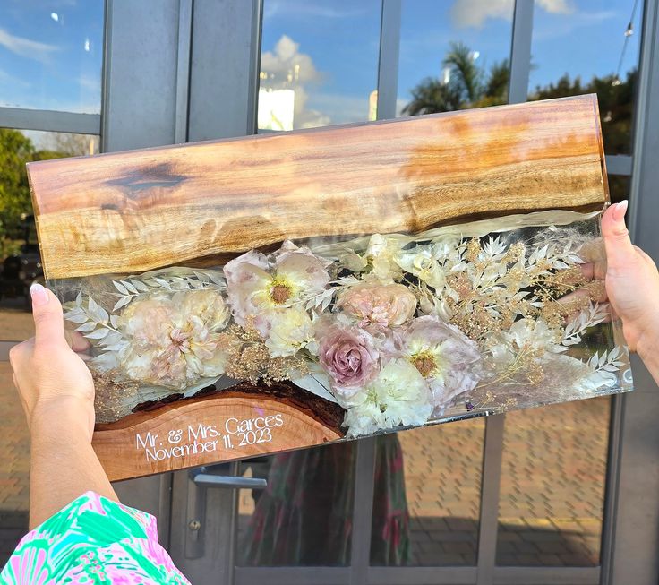 a person holding up a piece of wood with flowers on it in front of a building