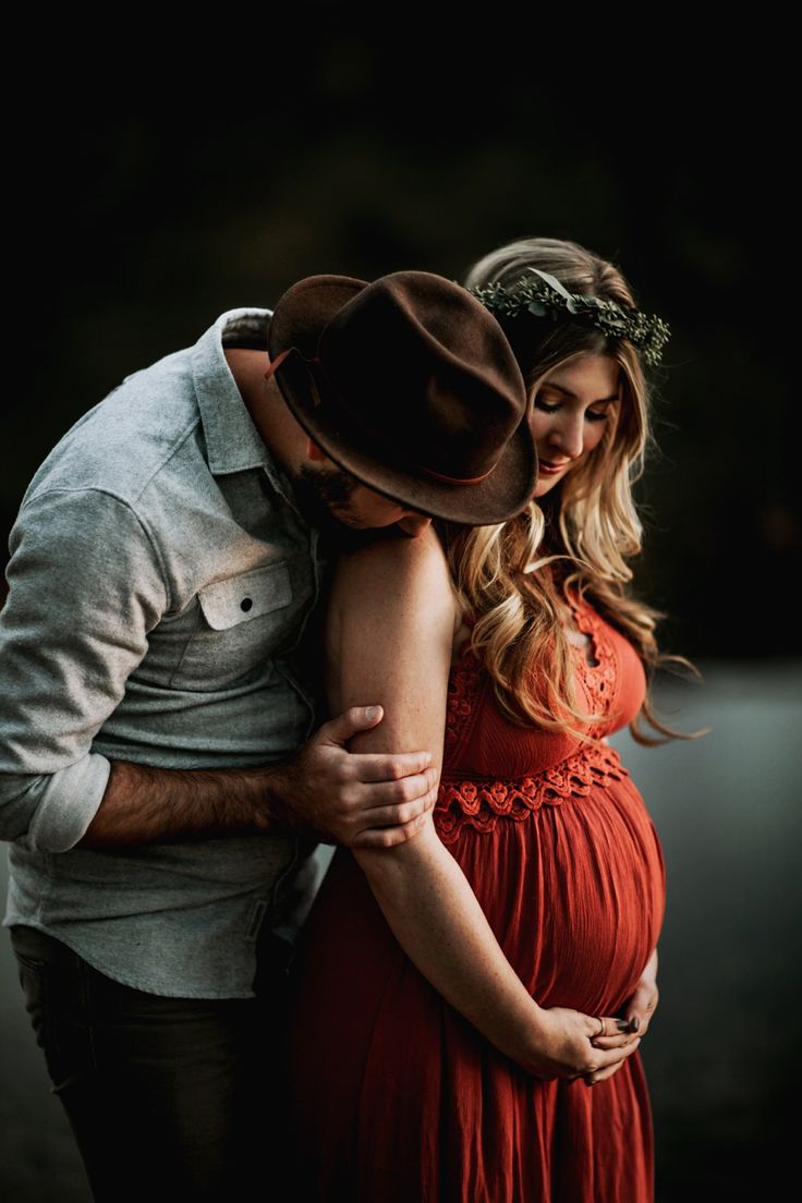 a pregnant woman in a red dress and cowboy hat hugs her husband's belly