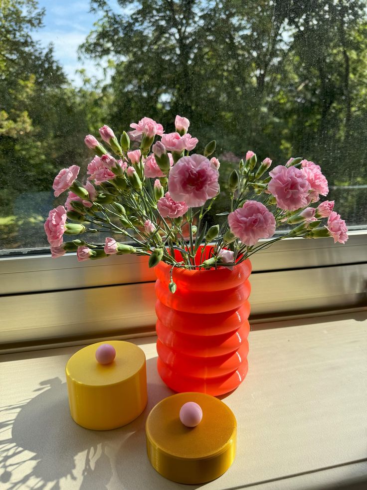 pink flowers are in a red vase next to yellow containers on a window sill