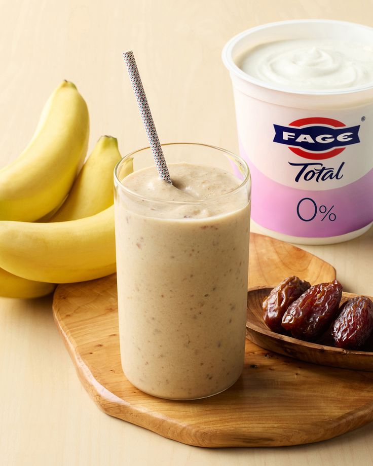 a smoothie in a glass next to bananas and yogurt on a wooden tray