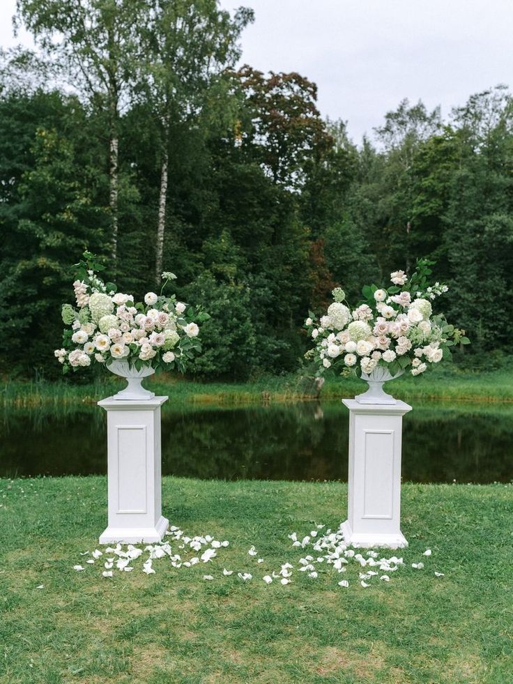 two white pedestals with flowers on them in front of a pond and trees behind them