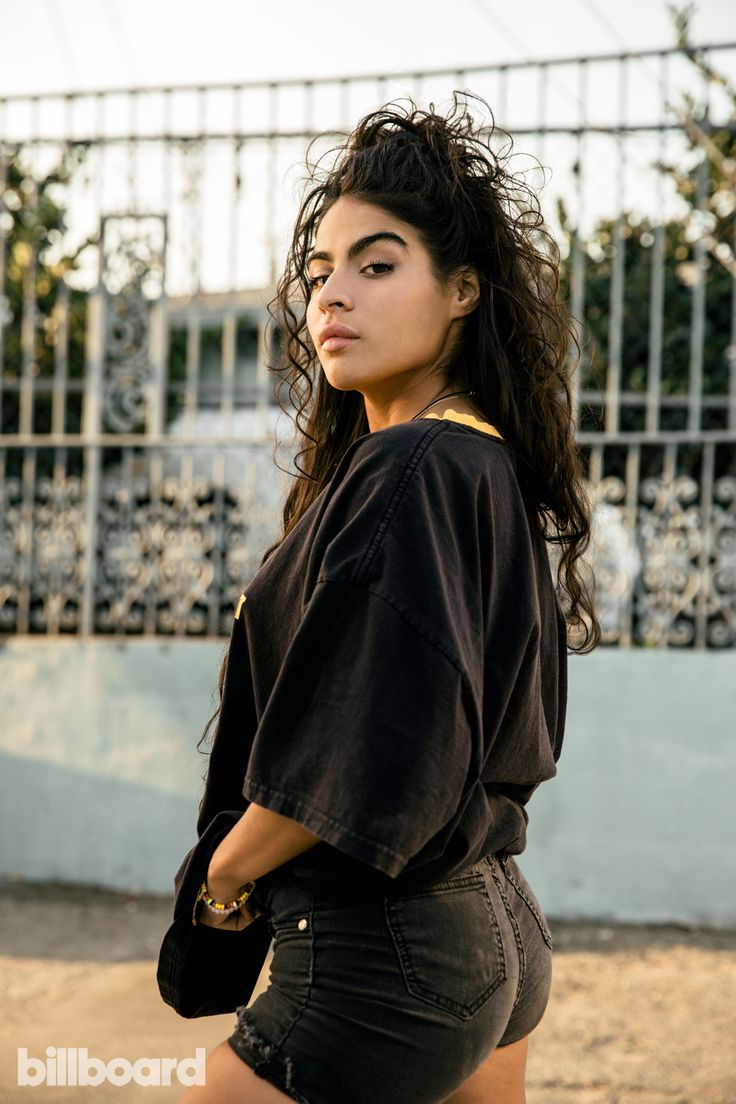 a woman with long dark hair is standing in front of a fence and looking at the camera