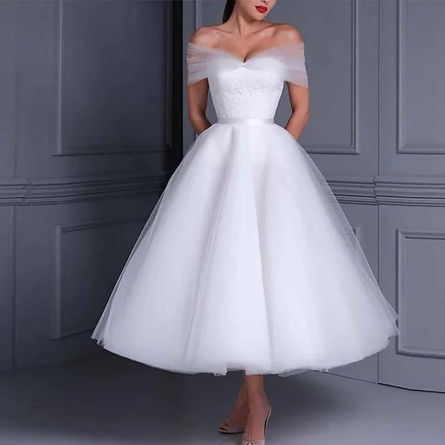 a woman wearing a white wedding dress standing in front of a gray wall with her hands on her hips