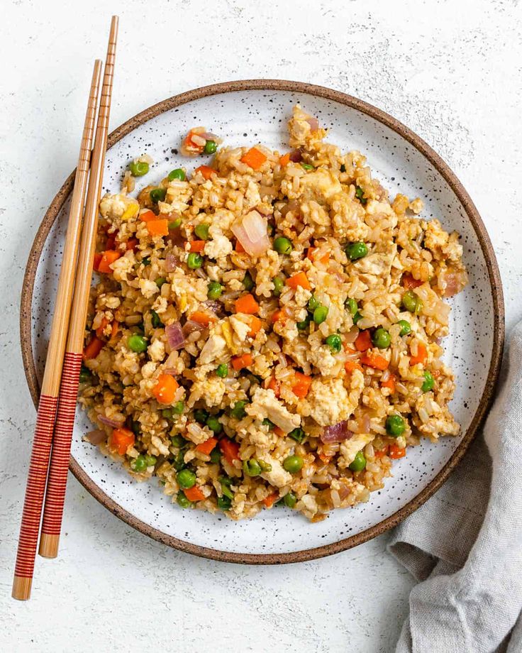 a plate full of rice and vegetables with chopsticks next to it on a white surface