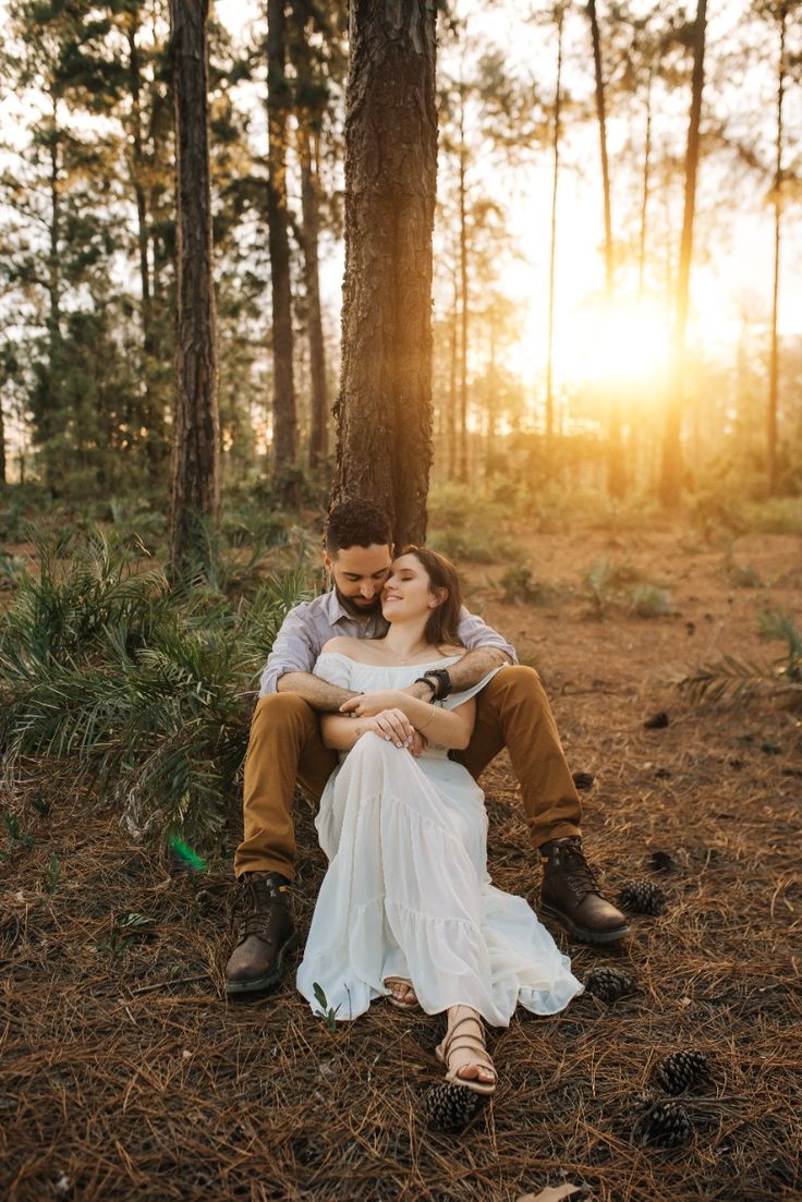 a man and woman are sitting on the ground in front of some trees at sunset