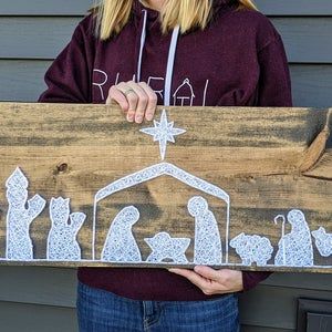 a woman holding up a wooden sign with the nativity scene painted on it's side