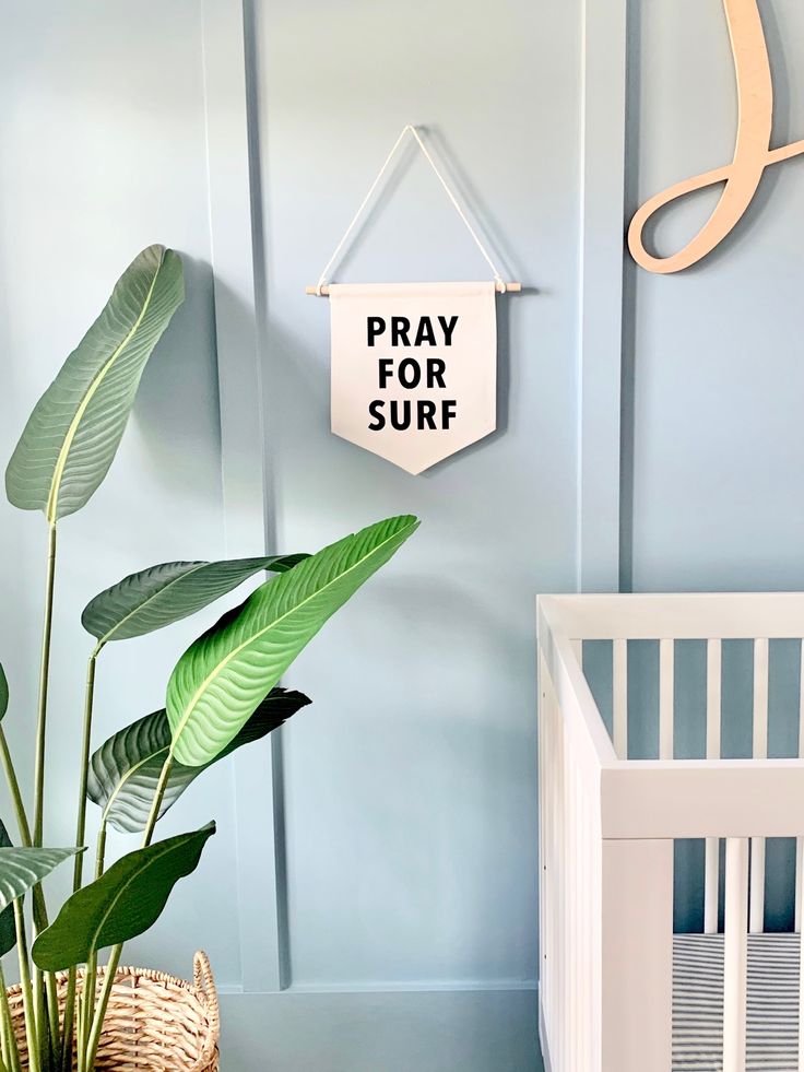 a white crib with a potted plant next to it and a sign that says pray for surf hanging on the wall