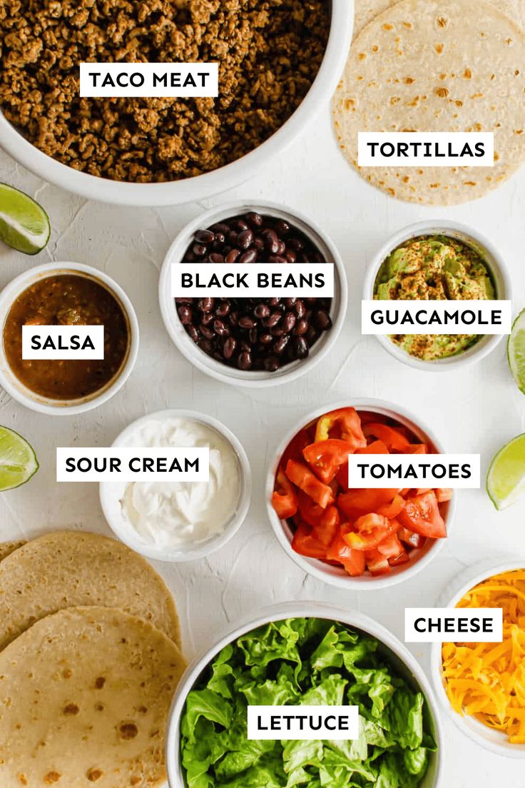 the ingredients for taco salad laid out in bowls on a white surface with limes, tortillas, black beans, sour cream, lettuce, and more