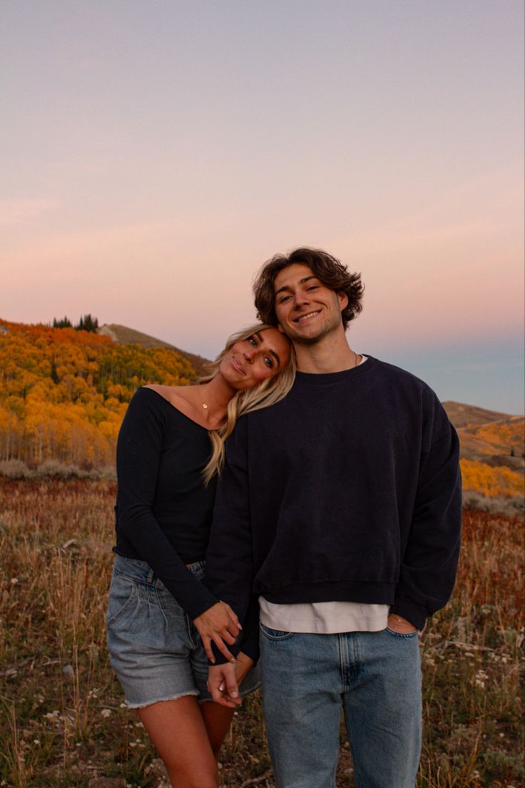 a man and woman standing next to each other in a field with trees behind them