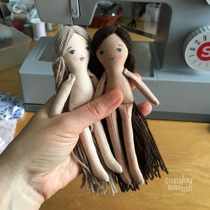 a hand holding two small dolls in front of a sewing machine