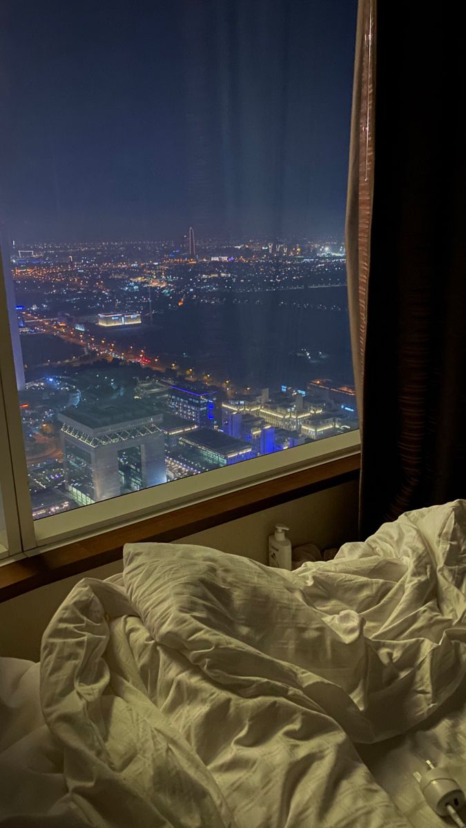 an unmade bed in front of a large window overlooking the city lights at night