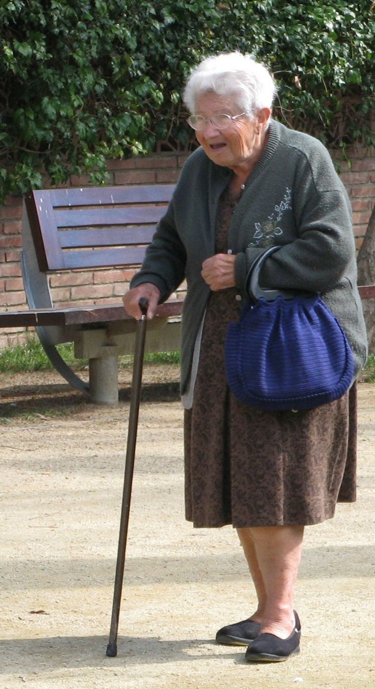 an old woman with a cane standing in front of a bench and holding a purse