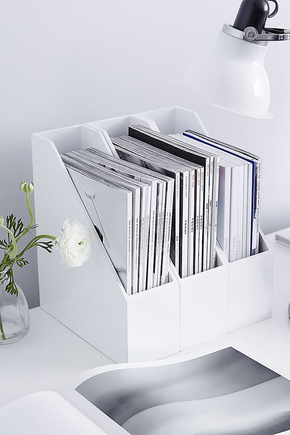 a white desk with a bunch of magazines and flowers in a vase next to it