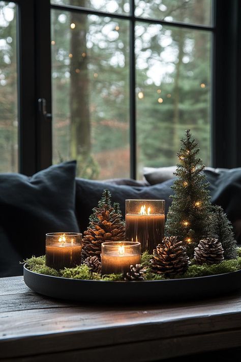 candles are lit on a tray with pine cones and evergreens in front of a window