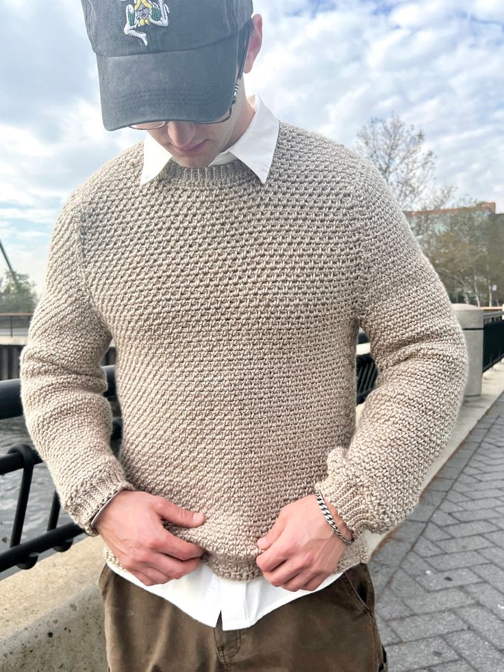 a man wearing a hat and sweater standing next to a fence with his hands in his pockets