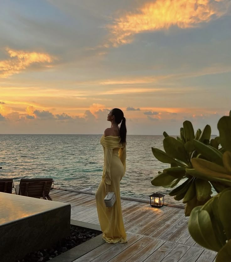 a woman in a yellow dress is standing on a dock by the ocean at sunset