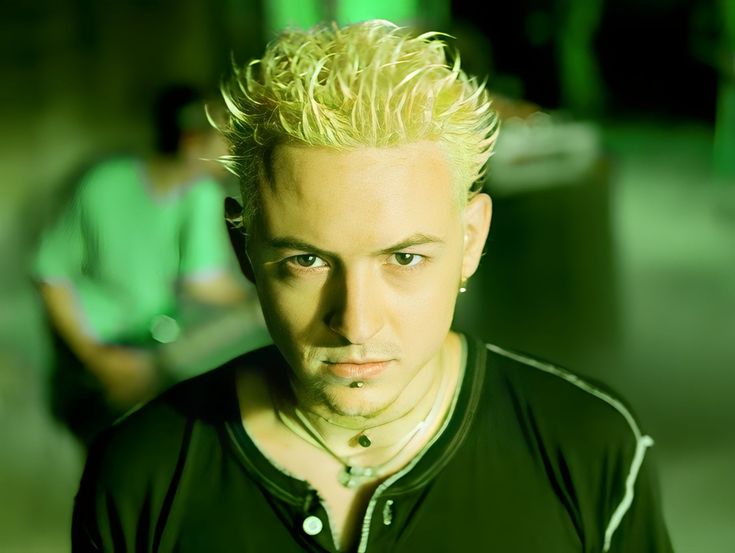 a man with blonde hair and piercings on his face looking at the camera while another person sits in the background