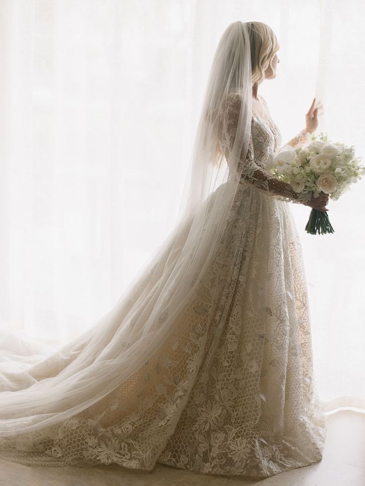 a woman in a wedding dress holding a bouquet and looking out the window at something