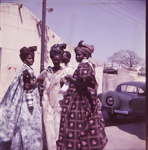 three women in dresses standing next to each other near a building and an old car