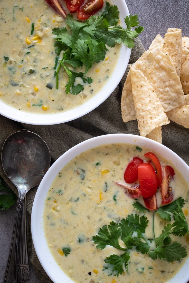 two white bowls filled with soup next to tortilla chips and tomatoes on a table