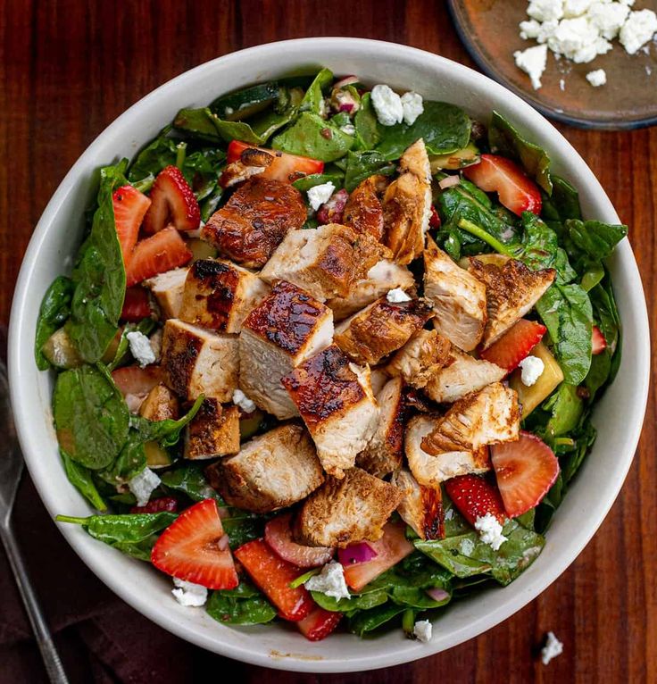 a salad with chicken, strawberries and feta cheese in a white bowl on a wooden table