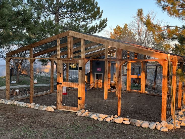 a chicken coop in the middle of a field with rocks and gravel around it, surrounded by pine trees