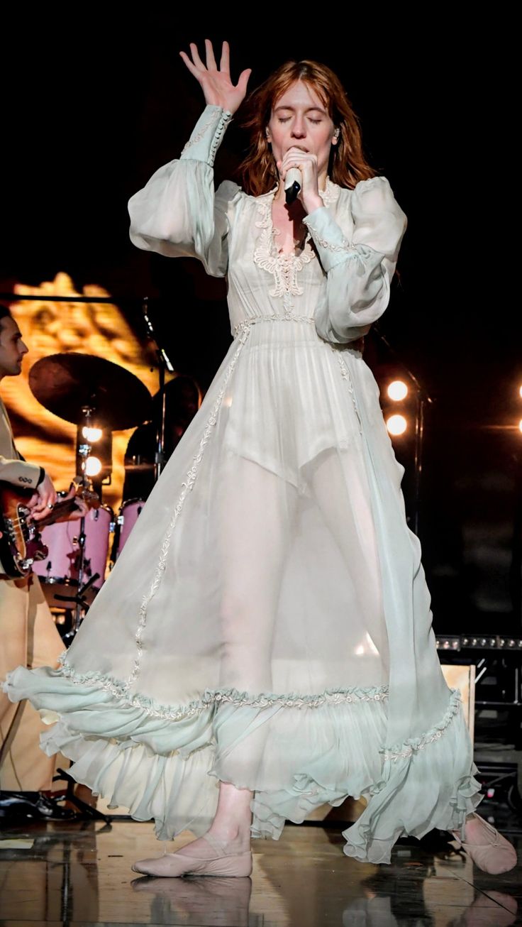 a woman in a white dress on stage with her hands up to the side and one hand raised