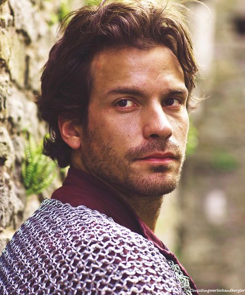 a man wearing a chain mail jacket leaning against a stone wall with his head turned to the side