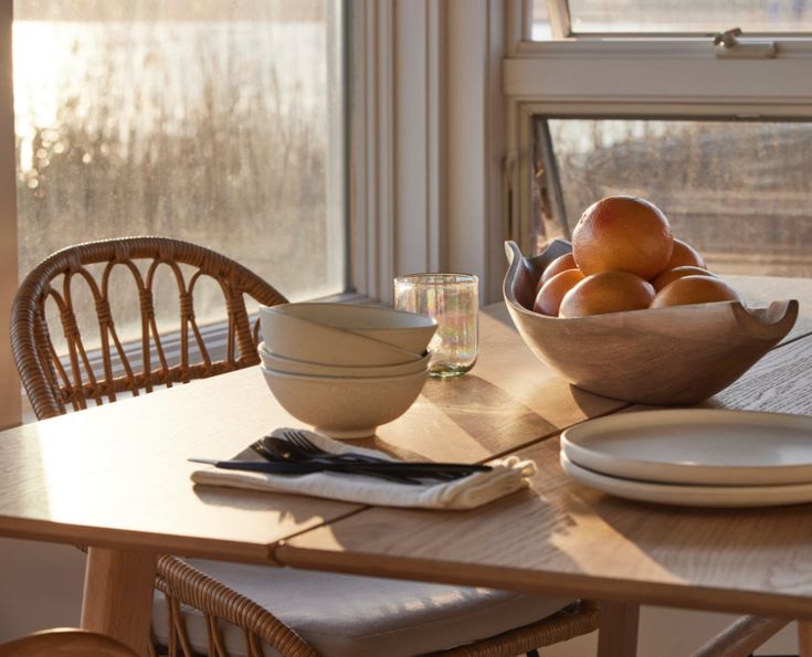 a bowl of oranges sitting on top of a wooden table next to a window