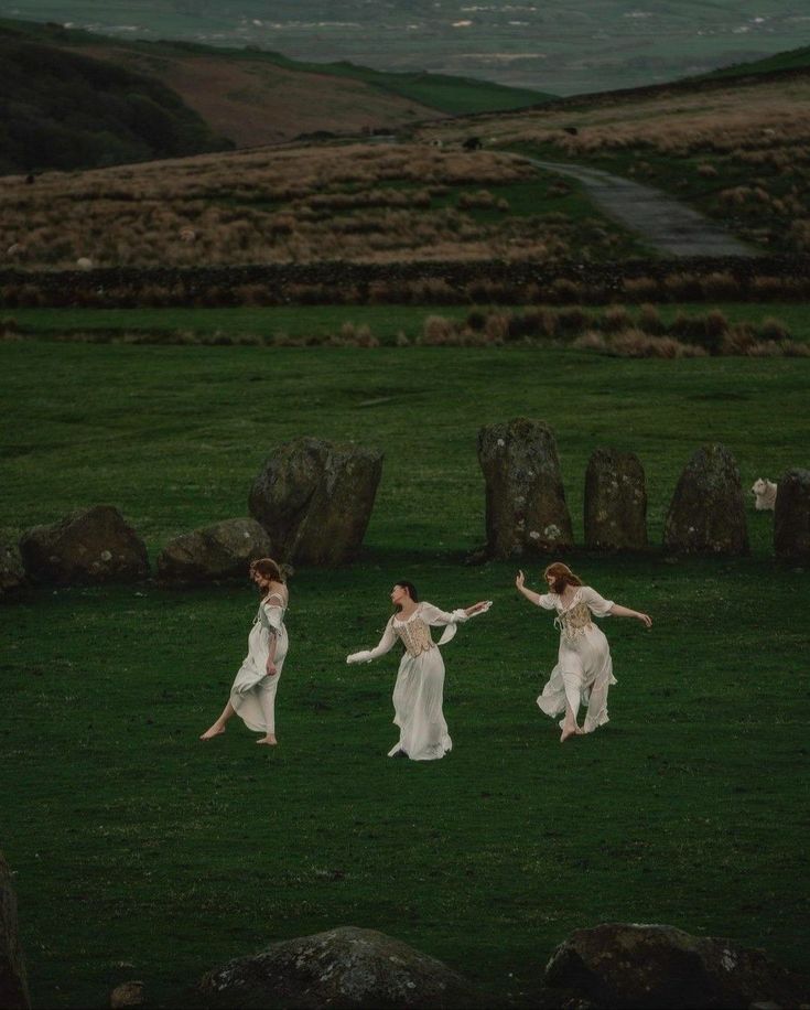 three women in white dresses are running through the grass with their arms spread out to each other