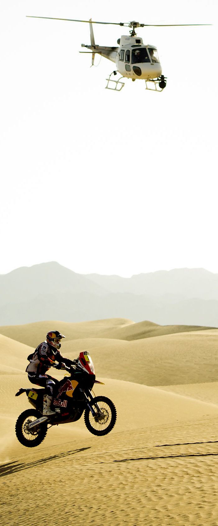 a person riding a motorcycle in the desert with a helicopter flying over them and a man on a motorbike behind him