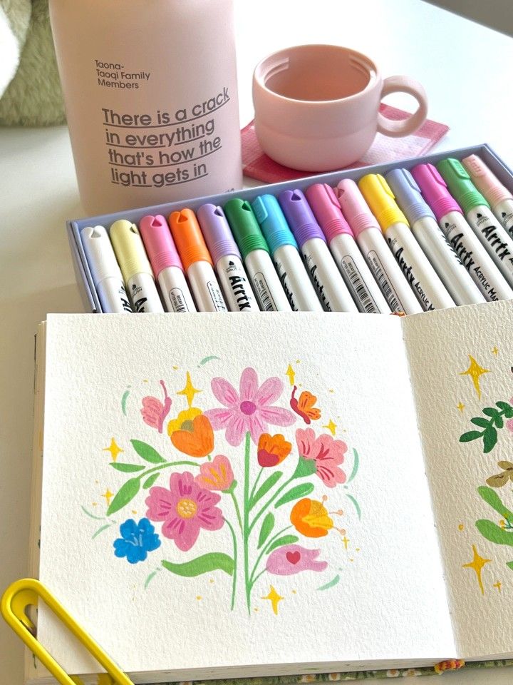 an open notebook with markers and pens next to it on a white table near a cup