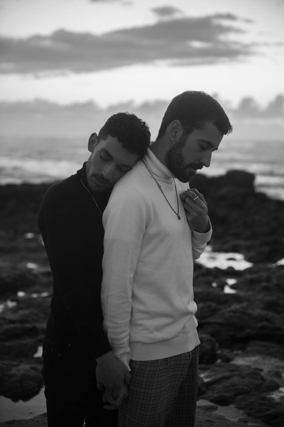 two men standing next to each other near the ocean with rocks and water in the background