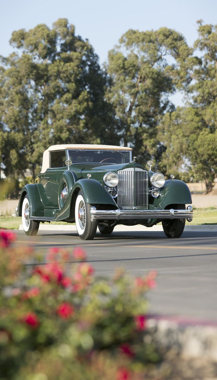 an old green car driving down the road in front of some bushes and trees with red flowers