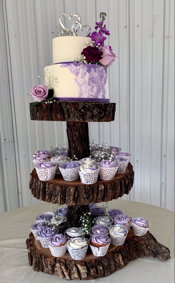 three tiered cake with cupcakes and flowers on the top layer, sitting on a tree stump