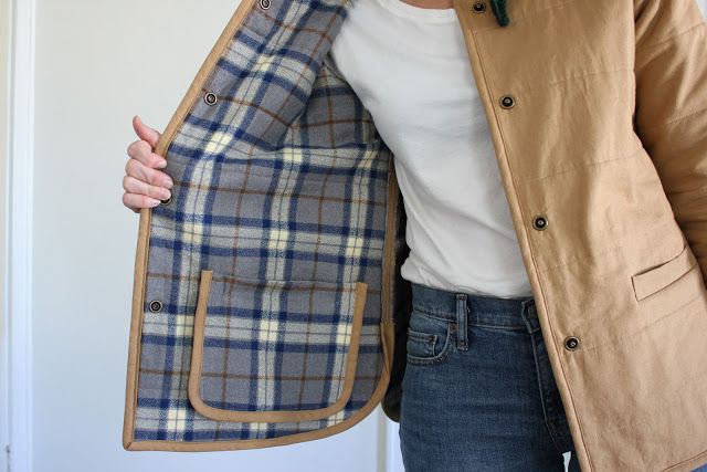 a woman wearing a tan jacket and blue jeans holds up a plaid coat over her shoulder