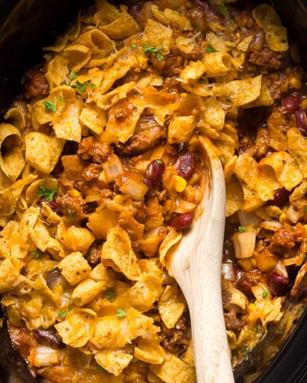 a crock pot filled with pasta, beans and meat in a red sauce next to a wooden spoon